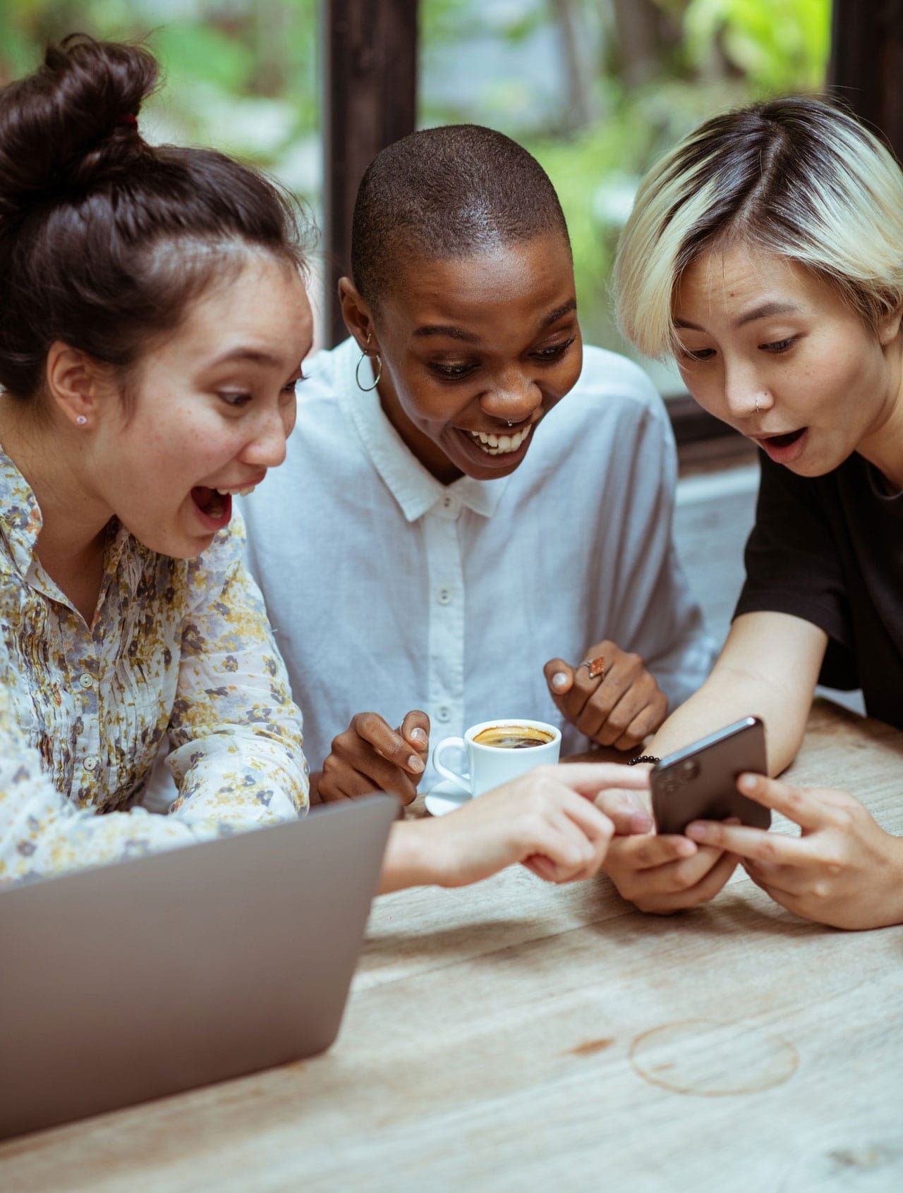 Três mulheres sentadas em volta de uma mesa olhando e mexendo para um celular, desmostrando felicidade e curiosidade.